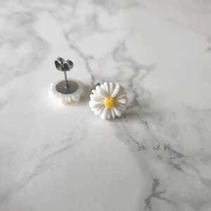 May include: A pair of silver stud earrings with white daisy flower accents. The center of each flower has a yellow circle.