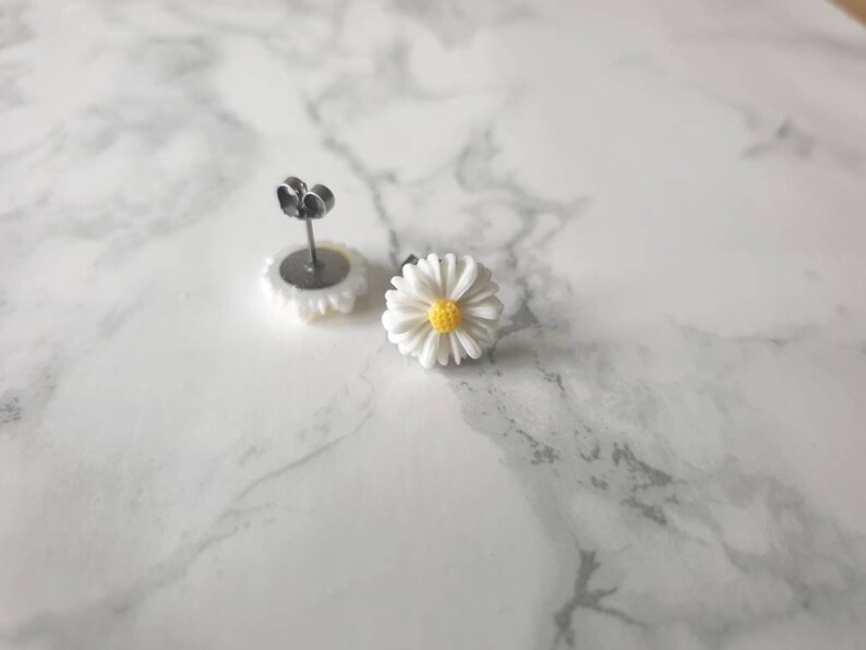 May include: A pair of silver stud earrings with white daisy flower accents. The center of each flower has a yellow circle.