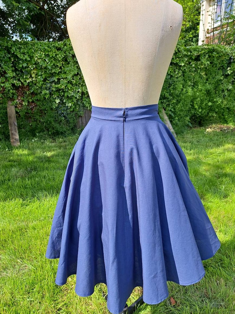 May include: A full-length view of a blue midi skirt with a high waist and a full circle shape. The skirt is made of a smooth, lightweight fabric and has a zipper closure in the back.