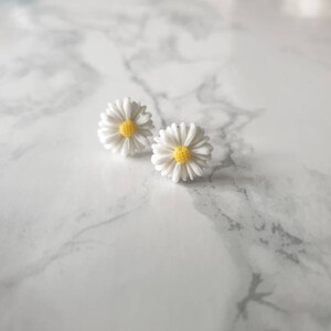 May include: A pair of white daisy earrings with yellow centers. The earrings are on a white marble surface.