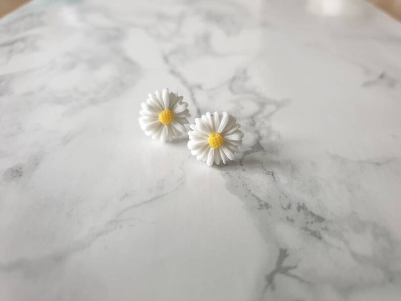 May include: A pair of white daisy earrings with yellow centers. The earrings are on a white marble surface.