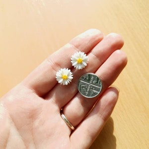 May include: Two small white daisy earrings with yellow centers, shown next to a British one pence coin for size comparison.