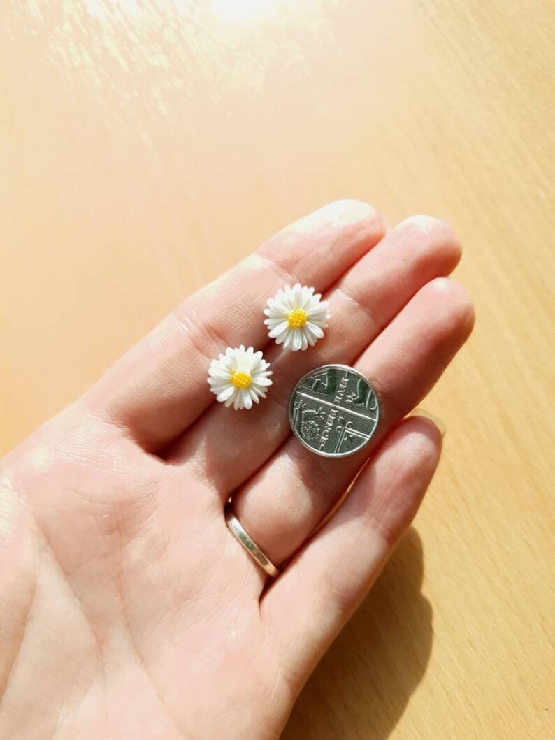 May include: Two small white daisy earrings with yellow centers, shown next to a British one pence coin for size comparison.