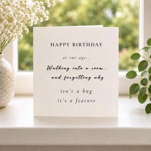May include: A square off-white greeting card with black text that reads: HAPPY BIRTHDAY at our age... Walking into a room... and forgetting why isn't a bug it's a feature. The card is positioned upright on a flat surface.