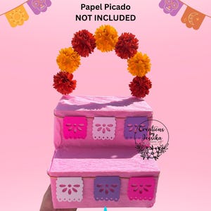 Puede incluir: Un expositor rosa de varios niveles decorado con papel picado colorido y una guirnalda de flores de papel naranja y rojo. El texto de la imagen dice "Completed Sample Flowers & Papel Picado NOT INCLUDED."