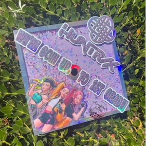 May include: A graduation cap topper with the text "WE'RE GOIN UP TO 1ST GRADE!" and "HUNTXX" in silver glitter. The topper features an illustration of three girls and a decorative knot design. The background is a light purple with glitter.