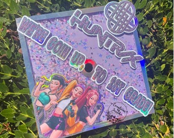 Puede incluir: Un adorno para gorra de graduación con el texto "WE'RE GOIN UP TO 1ST GRADE!" y "HUNTXX" en purpurina plateada. El adorno presenta una ilustración de tres chicas y un diseño de nudo decorativo. El fondo es de color lila claro con purpurina.
