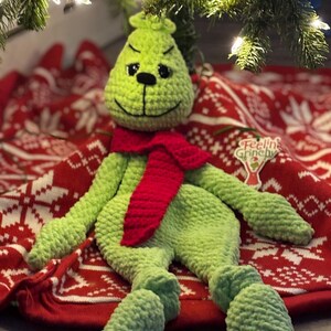 May include: A handmade, light green crocheted Grinch doll with a red scarf. The doll is lying on a red and white patterned Christmas tree skirt under a Christmas tree. The doll has black eyes and a stitched smile. The text "Feelin' Grinchy" is on the tree skirt.