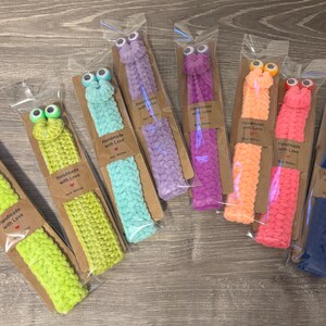 May include: A collection of handmade, colorful crochet bookmarks, each with googly eyes. The bookmarks are in various colors, including green, blue, purple, orange, and pink. Each bookmark is packaged with a small brown tag that reads "Handmade with Love."