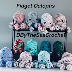 May include: A collection of handmade crochet fidget octopuses in various colors, including pink, blue, green, and rainbow. The octopuses are displayed on a two-tiered black metal shelf. The text "Fidget Octopus" and "DByTheSeaCrochet" are visible.