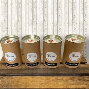 May include: Four cylindrical brown paper candle containers with silver lids are arranged on a wooden surface. Each container has a white label with a tree logo and a black text. The lids feature a Union Jack design.