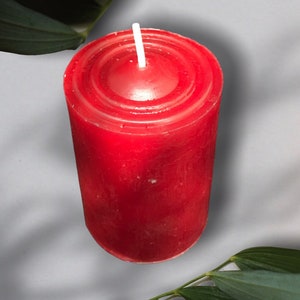 May include: A red pillar candle with a white wick. The candle is sitting on a white surface with green leaves in the background.