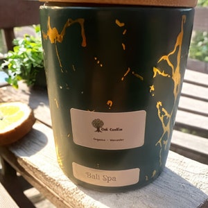 Pode incluir: Um frasco de vela verde com tampa de madeira e detalhes dourados. O frasco tem um rótulo que diz "Oak Candles" e "Organics - Worcester". A parte inferior do frasco diz "Bali Spa".