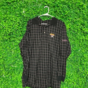 May include: Black and white plaid long-sleeve shirt with a red and yellow embroidered Pittsburgh Pirates logo. The shirt has a button-down collar and a single button closure.