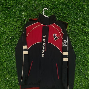 以下が含まれることがあります： 黒と赤のヒューストン・テキサンズのジャケット。袖に白いストライプ、胸にチームロゴが入っています。ジッパーとフードが付いています。