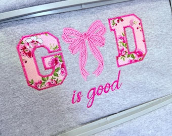 Könnte beinhalten: Grauer Stoff mit den gestickten Worten "GOD is good". Die Buchstaben "G" und "D" sind rosa mit einem Blumenmuster, und die Schleife ist rosa. Die Stickerei befindet sich in einem Rahmen.