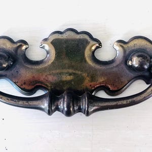 Puede incluir: Un tirador de cajón metálico con un acabado oscuro y deslustrado. El herraje presenta una placa decorativa ancha con bordes curvos y festoneados con una cresta central, junto a un asa arqueada oscilante con un detalle redondeado.