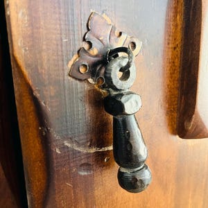 May include: A dark metal drop handle with a hammered texture attached to a wooden surface by a decorative, ornate backplate. The handle features a ring-style attachment point and a tapered, cylindrical body with a rounded, bulbous end.