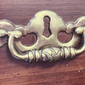 May include: A brass drawer pull featuring a decorative backplate with a central keyhole cutout and a swinging bail handle. The handle is adorned with a detailed embossed wheat stalk motif at its center, set against a dark wood surface.