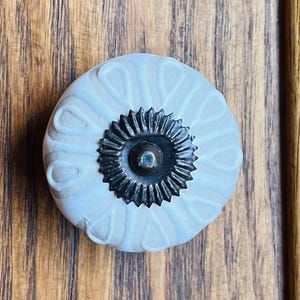 Peut inclure: Un bouton de placard rond en céramique bleu clair présentant un motif en relief en forme de pétales sur toute sa surface. Le centre du bouton est orné d'une plaque décorative métallique sombre en forme de soleil avec une petite saillie centrale.