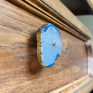 May include: A drawer knob featuring a slice of light blue agate stone with a natural, uneven edge finished in metallic gold. The stone has a smooth, translucent surface with subtle banding and is attached to a wooden drawer front with a metal screw.