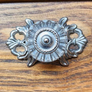 Può includere: Un pomello per cassetto con un centro in vetro trasparente sfaccettato, un bordo circolare in metallo argentato e un punto centrale. Il vetro è montato su una placca posteriore in metallo argentato ornata con elementi decorativi curvi a forma di foglia.