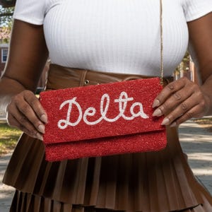 May include: A red beaded clutch with the word "Delta" in white script. The clutch has a gold chain strap and a rectangular shape. The background is blurred, but appears to be outdoors.