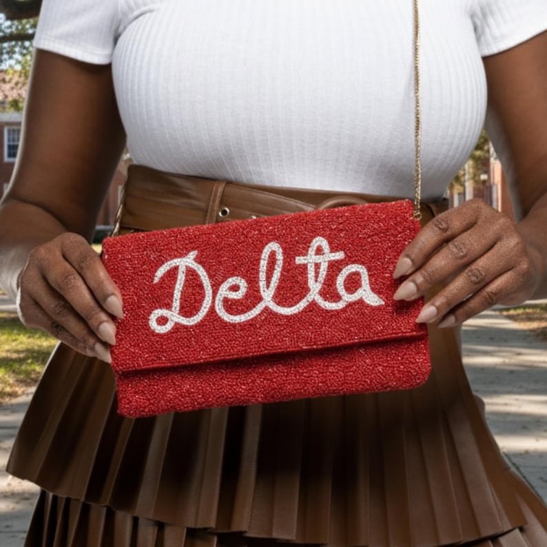 May include: A red beaded clutch with the word "Delta" in white script. The clutch has a gold chain strap and a rectangular shape. The background is blurred, but appears to be outdoors.