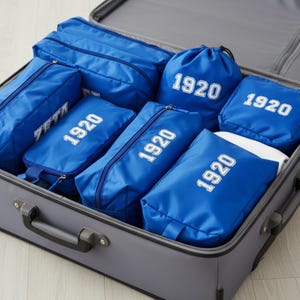 May include: A gray suitcase filled with blue packing cubes and bags. The bags have white text that reads "1920" and "ZETA". The suitcase is open, revealing the organized contents, perfect for travel.
