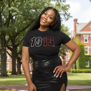 May include: A black t-shirt with the year "1913" in white and red rhinestones. The shirt is paired with a black, shiny skirt. The image is taken outdoors with a background of trees and a brick building.