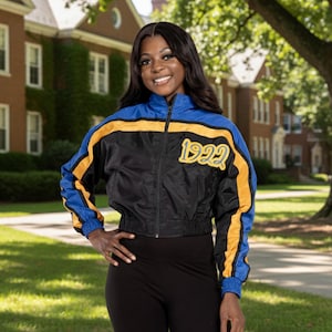 May include: Black, blue, and gold cropped jacket with the year "1922" printed on the front. The jacket has a zipper closure and a sporty design. The sleeves and shoulders are blue, with gold stripes. The jacket is worn with black leggings.