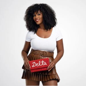 May include: A red beaded clutch with the word "Delta" in white script is held in front of a person. The clutch is rectangular and appears to be covered in small beads. The person is wearing a white top, a brown pleated skirt, and a brown belt.
