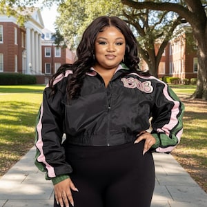 May include: Black bomber jacket with pink and green accents, featuring the year "1903" on the chest. The jacket has a zip-up front and is paired with black leggings. The sleeves have green and pink stripes.