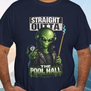 May include: A navy blue t-shirt featuring a graphic of a green alien wearing headphones and a black leather jacket. The alien holds an 8-ball and a pool cue with a blue flame tip. The text reads STRAIGHT OUTTA THE POOL HALL in white and green.