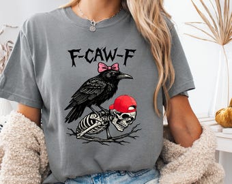 May include: Gray t-shirt featuring a black crow with a pink bow perched atop a skeleton wearing a red baseball cap. The text "F-CAW-F" is above the crow. The shirt is a casual, comfortable style.