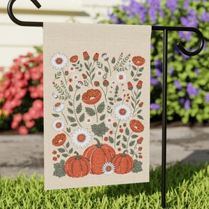 May include: A beige garden flag featuring a fall-themed design. The flag displays orange pumpkins, white daisies, and red poppies amidst green foliage. The flag is hanging on a black metal stand, perfect for outdoor decor.