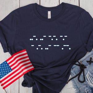 May include: Navy blue t-shirt featuring a Morse code design in white dots and dashes. An American flag is visible in the lower left corner. The shirt is paired with denim shorts.