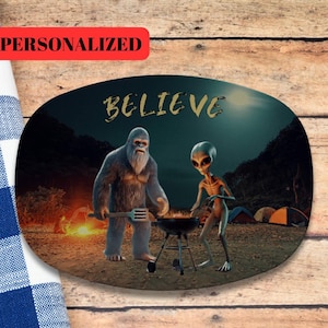 May include: Oval-shaped serving platter featuring a Bigfoot and an alien grilling over a barbecue. The word "BELIEVE" is written in gold lettering. The word "PERSONALIZED" is in a red rectangle at the top. Tents are in the background.