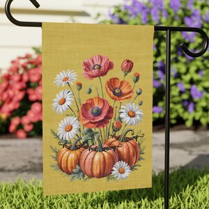 May include: A yellow garden flag with a floral design including poppies, daisies, and pumpkins. The pumpkins have the text "Fuck Trump". The flag is displayed on a black metal stand in a garden setting.