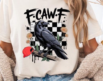 May include: A cream-colored t-shirt featuring a graphic print of a black raven perched on a branch. The design includes a black and white checkered square background, orange daisy flowers, a red baseball cap, and the text F.CAW.F in black.