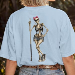 Peut inclure: Un t-shirt bleu clair avec une illustration de squelette portant un nœud rose, des lunettes de soleil et des créoles. Le squelette porte un short en jean avec des bretelles et des baskets noires, tenant une queue de billard avec deux boules 8.