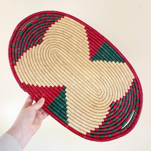 May include: Oval woven basket with a natural beige center and a red and green geometric pattern around the edges. The basket is held by a hand and is against a light background. The basket is approximately 16 inches long.