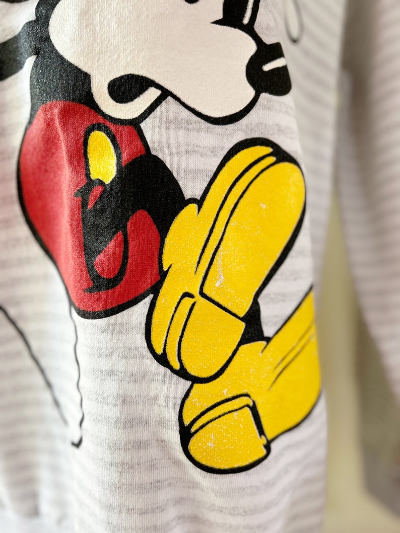 May include: A gray sweatshirt with a black and white cartoon image of Mickey Mouse. Mickey is wearing red shorts and yellow shoes. The sweatshirt has a striped pattern.