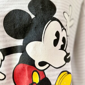 May include: A white and gray striped shirt with a black and white cartoon image of Mickey Mouse. Mickey Mouse is wearing red shorts and yellow shoes.
