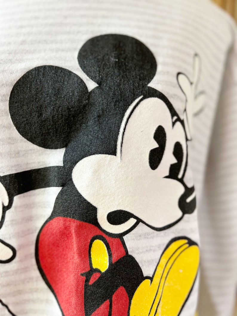 May include: A white and gray striped shirt with a black and white cartoon image of Mickey Mouse. Mickey Mouse is wearing red shorts and yellow shoes.