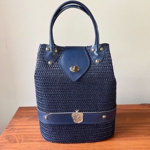 May include: A navy blue woven handbag with a leather strap and a silver lock. The bag has a decorative silver emblem on the front.