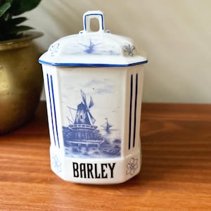 May include: White ceramic barley jar with a blue windmill and boat design. The jar has a lid with a handle and blue trim. The word "BARLEY" is printed in black on the front.