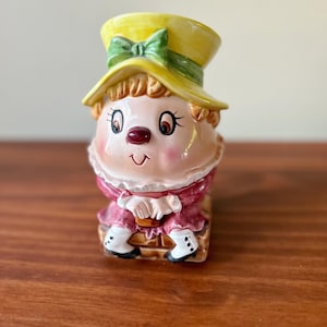 May include: A ceramic figurine of a girl wearing a yellow hat with a green bow and a pink dress. She is sitting on a brown brick.