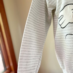 May include: A gray and white striped long-sleeve sweatshirt with a black and white cartoon hand design on the front.