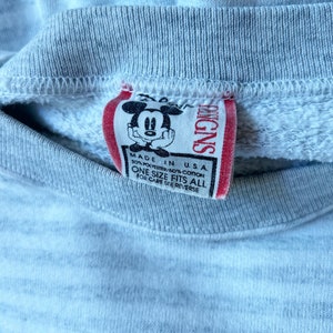 May include: A gray sweatshirt with a red tag featuring a cartoon Mickey Mouse. The tag reads "Disney" and "Made in USA 50% Polyester/50% Cotton One Size Fits All For Care See Reverse."